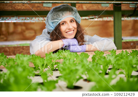 Happy woman agronomist checking plant growth in greenhouse. Joyful female gardener looking at green leafy plants and smiling while standing near shelf with seedlings. 109212429