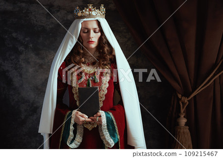 medieval queen in red dress with book, veil and crown 109215467