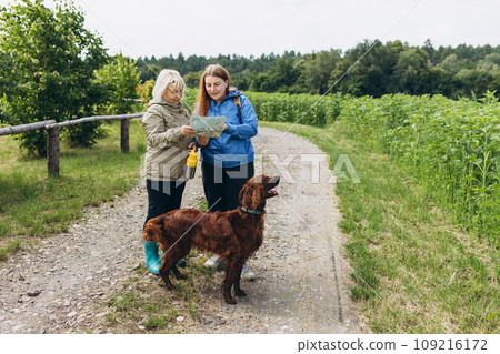 Two happy Womans in active trekking clothes with map having a halt after hiking. Travel and active lifestyle concept, full body. Travel and hiking with pet. 109216172
