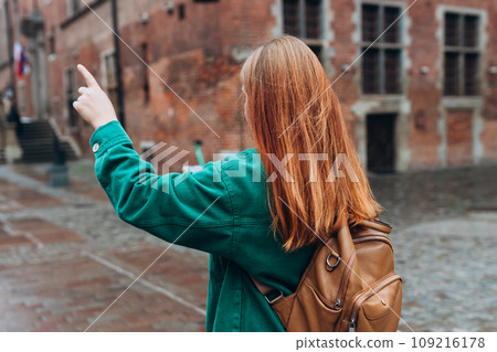 Casual dressed woman 30 years old exploring city streets during international excursion in town. Redhead girl with backpack pointing finger on city street in Gdansk. Traveling Europe in autumn. 109216178