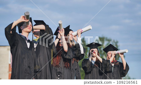 Cheerful graduates on a sunny day look through diplomas like a telescope. 109216561