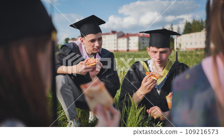 Graduates in black suits eating pizza in a city meadow. Graduates in black suits eating pizza in a city meadow. 109216573