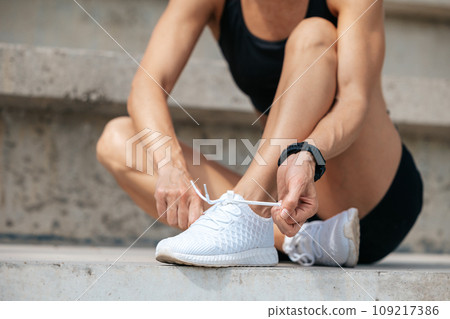 Female kneeling and tying shoelace on street on sunny day. Female kneeling and tying shoelace on street on sunny day. 109217386