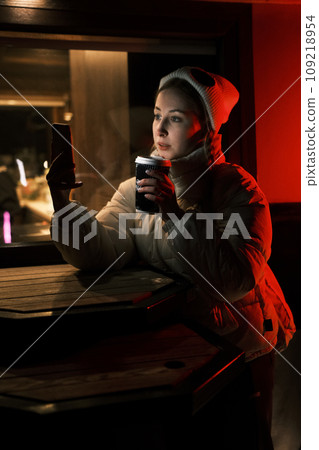 Model blonde with tails with cup of coffee near coffee shop with red and yellow light looking in smartphone. Girl weared white dress, jacket and hat posing near cafe in winter night 109218954