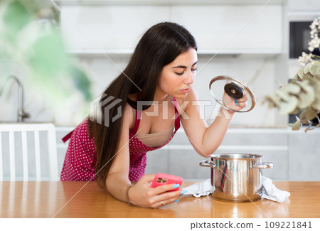 Young woman holding mobile phone opening lid of the pan in the kitchen 109221841