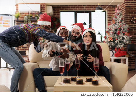Happy coworkers drinking alcohol and enjoying console gaming at christmas party in decorated office. Cheerful colleagues celebrating new year winter seasonal holiday together 109222009