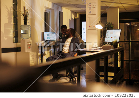 Smiling businessmen riding desk chair having fun during moving activity in startup office. Cheerful employees taking break from work enjoying doing sport races late at night. Business concept Smiling businessmen riding desk chair having fun during moving activity in startup office. Cheerful employees taking break from work enjoying doing sport races late at night. Business concept 109222023