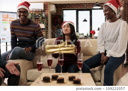 Asian woman worker receiving present from coworkers while celebrating christmas in decorated office. Company employee sitting on couch with xmas gift box at corporate holiday party 109222057