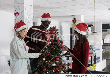 Colleagues decorating Christmas tree with ornaments and lights garland at work in xmas adorn workspace. Staff members enjoying time spent together during winter holiday season 109222087
