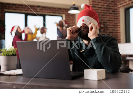 Frustrated tired employee putting santa hat on eyes while working overtime during christmas season in office. Startup company worker managing tasks workload during winter holiday 109222122