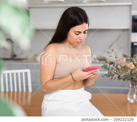 Young woman sitting on kitchen table and looking at her phone 109222126
