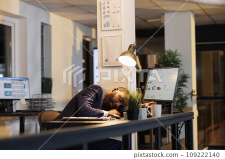 Businessman napping on desk because of overwork in startup office. Workaholic executive manager falling asleep after working late at night at company investment plan. Business concept Businessman napping on desk because of overwork in startup office. Workaholic executive manager falling asleep after working late at night at company investment plan. Business concept 109222140