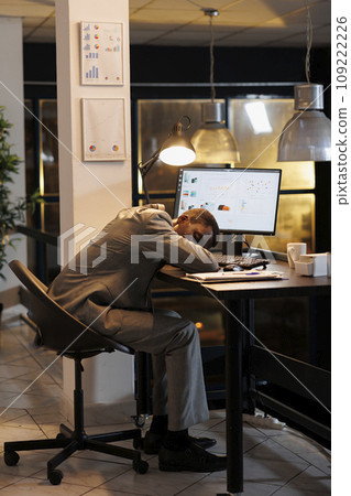 Exhausted overworked executive manager sleeping on desk table in startup office after planning company investment plan late at night. Workaholic entrepreneur working at marketing project. Exhausted overworked executive manager sleeping on desk table in startup office after planning company investment plan late at night. Workaholic entrepreneur working at marketing project. 109222226