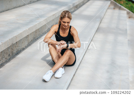 Female runner checking results on smart watches sitting on stairs taking pulse after jogging. 109224202
