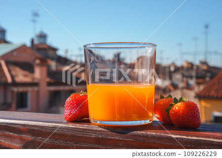 A refreshing glass of orange juice with strawberries sits on a neutral-toned balcony. This image is part of a collection showcasing popular places in Venice, Italy. A refreshing glass of orange juice with strawberries sits on a neutral-toned balcony. This image is part of a collection showcasing popular places in Venice, Italy. 109226229