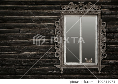 upper half of a tabby cat's muzzle looking out of a antique wooden carved window on an old log wall upper half of a tabby cat's muzzle looking out of a antique wooden carved window on an old log wall 109234973