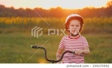 Young girl in helmet on head stands laughing with bike on field at sunset Young girl in helmet on head stands laughing with bike on field at sunset 109235692