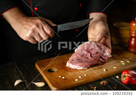 The cook cuts beef meat with a knife in the kitchen, prepares food. Peppers and spices on the kitchen table to prepare a delicious lunch. The cook cuts beef meat with a knife in the kitchen, prepares food. Peppers and spices on the kitchen table to prepare a delicious lunch. 109238382