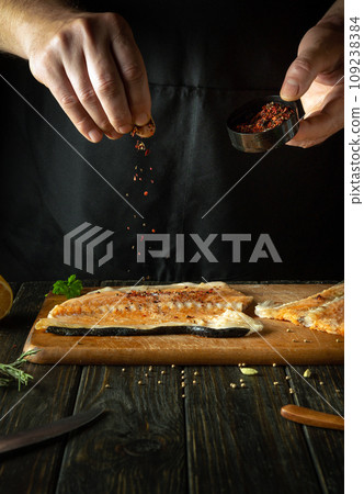 Adding dry spices to fish steaks for aroma and taste. American cuisine. The process of preparing fish by the hands of a chef in a restaurant or hotel kitchen Adding dry spices to fish steaks for aroma and taste. American cuisine. The process of preparing fish by the hands of a chef in a restaurant or hotel kitchen 109238384