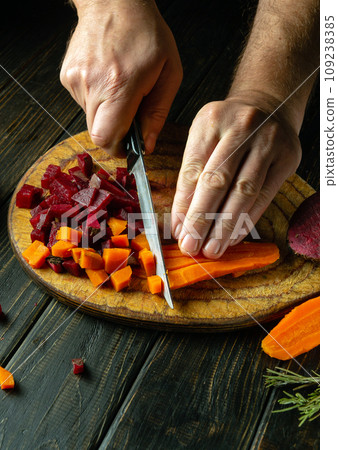 The cook prepares a vinaigrette from boiled vegetables. Slicing carrots and beets with a knife on a wooden cutting board 109238385