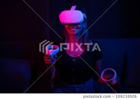 Young asian woman using at home Virtual Reality headset with joysticks playing VR video games, neon pink blue light Young asian woman using at home Virtual Reality headset with joysticks playing VR video games, neon pink blue light 109238926