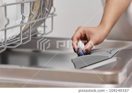 female hand puts dishwasher tablet into open automatic built-in dishwasher machine with dirty dish inside. Close-up female hand puts dishwasher tablet into open automatic built-in dishwasher machine with dirty dish inside. Close-up 109240387