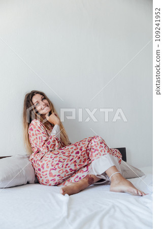 Photo of young happy woman in pajama stretching her arms and smiling while sitting on bed after sleep or nap 109241952