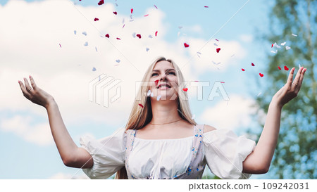 A girl of blonde Slavic appearance and white dress tosses confetti of hearts in summer. A girl of blonde Slavic appearance and white dress tosses confetti of hearts in summer. 109242031