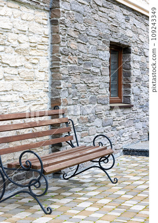 A bench with wrought-iron railings against the background of a textured stone wall of a house in the daytime. A bench with wrought-iron railings against the background of a textured stone wall of a house in the daytime. 109243349