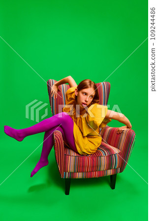 Young attractive girl in unusual, strange, variegated colored outfit and posing extraordinary in retro chair against green background. 109244486