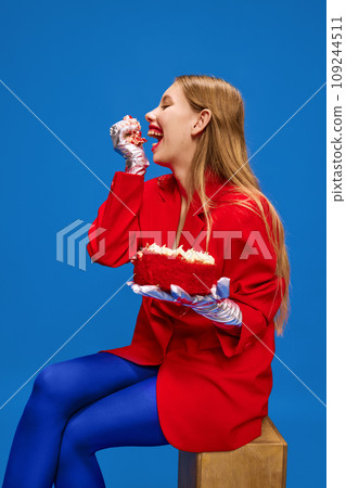 Portrait of overjoyed, unusual, vivid dressed young girl sitting and eating broken off sweet and tasty big piece of birthday cake against blue background. 109244511
