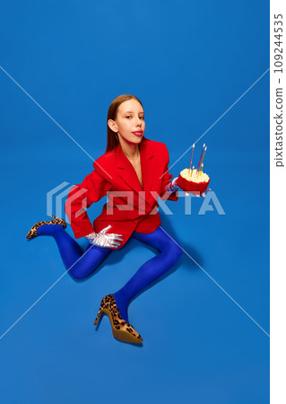 Young lady dressed unusual, freaky, bright outfit, wearing blue tights, red jacket and animal printed heels holding birthday cake with candles. Young lady dressed unusual, freaky, bright outfit, wearing blue tights, red jacket and animal printed heels holding birthday cake with candles. 109244535