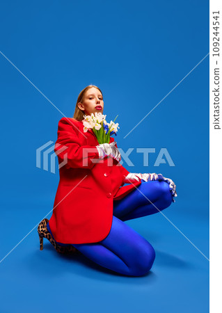 Weird girl in blue tights posing with flowers in unusual, strange, red jacket sitting against blue color studio background. Weird girl in blue tights posing with flowers in unusual, strange, red jacket sitting against blue color studio background. 109244541