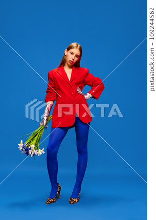 Full-length portrait of young girl in unusual, strange, variegated colored outfit and posing with flower bunch against color studio background. 109244562