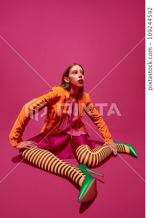 Full-length portrait of expressive young girl, lady in unusual, variegated orange striped tights, long skirt and short jacket against magenta background. Full-length portrait of expressive young girl, lady in unusual, variegated orange striped tights, long skirt and short jacket against magenta background. 109244592
