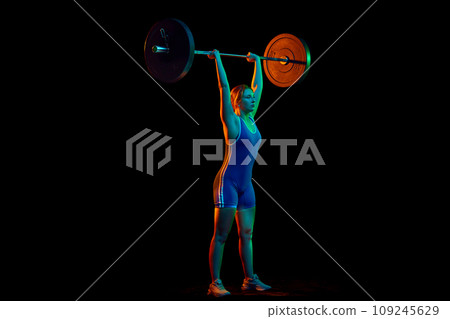 Champion. Strong young girl, athlete in sportswear lifting heavy weight, barbell against black background in neon light 109245629