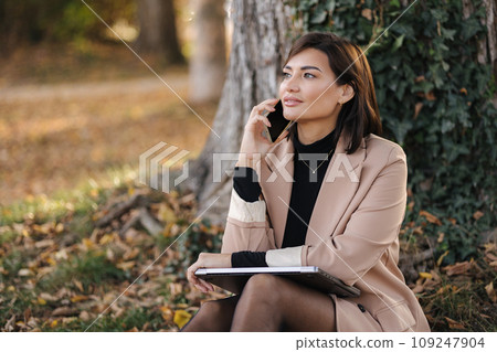 Portrait of attractive female sitting in the park and speak by the phone. Happy woman spent time on open air 109247904