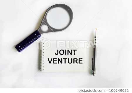joint venture. text on a notepad on a white background next to a magnifying glass 109249021