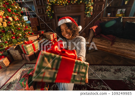 Merry Christmas. African American woman giving you present gift box near Christmas tree. Girl in living room with Christmas tree and fireplace packing wrapping opening gift box. Christmas eve at home 109249985