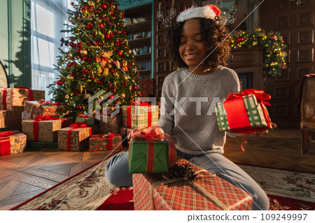 Merry Christmas. African American woman packing wrapping gift box near Christmas tree. Girl in living room with Christmas tree and fireplace opening gift box. Christmas eve at home 109249997