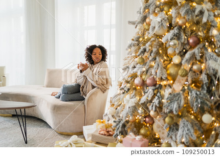 Merry Christmas. African American woman with cup of hot drink coffee tea sitting near Christmas tree at home. Girl in living room with Christmas tree. Christmas hygge holidays at home 109250013