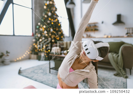 Practicing yoga poses, a fit young woman wears virtual reality glasses next to a Christmas tree. 109250432