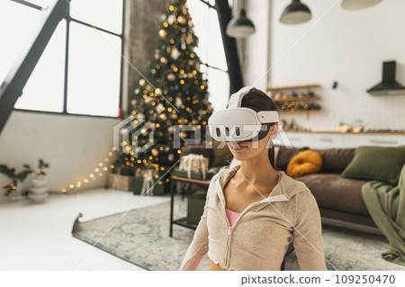 Demonstrating yoga poses, a young woman uses virtual reality glasses near a Christmas tree. 109250470