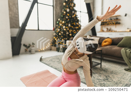Engaging in yoga poses, a sporty young lady wears VR glasses beside a Christmas tree. 109250471