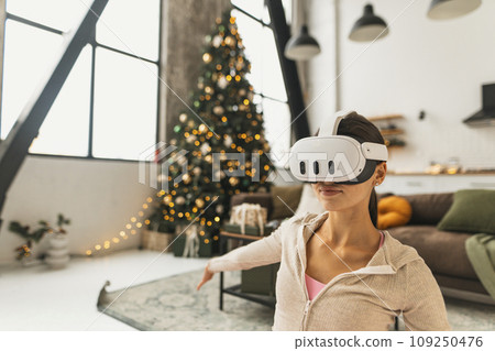 A fitness-oriented young woman practices yoga poses while wearing VR glasses by a Christmas tree. A fitness-oriented young woman practices yoga poses while wearing VR glasses by a Christmas tree. 109250476