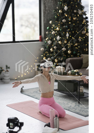 The stylish lady, dressed in sportswear and with a virtual reality headset, practices yoga near a decorated Christmas tree. 109250493