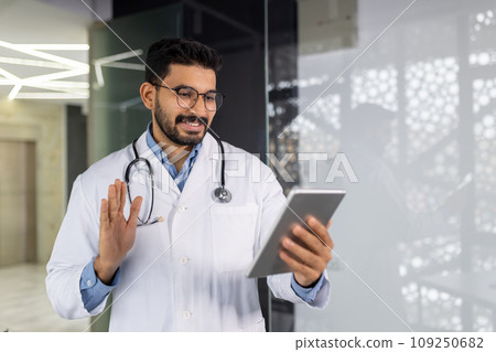 A young doctor smiles with a tablet computer in his hands, talks remotely with a patient, consults using a video game, waves and greets, stands inside the clinic by the window. A young doctor smiles with a tablet computer in his hands, talks remotely with a patient, consults using a video game, waves and greets, stands inside the clinic by the window. 109250682