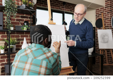 People connecting while learning new skills, two diverse men looking at easel with canvas discussing artwork while studying pencil drawing together at group art class. Sketching lesson 109252399