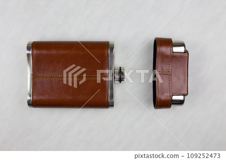 Brown leather flask for alcohol lies next to an open lid on a white table, top view Brown leather flask for alcohol lies next to an open lid on a white table, top view 109252473