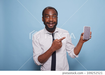 Enthusiastic black man pointing at smartphone blank screen showing mobile feature of mobile game or app. Excited individual using index finger to enhance features of digital device. 109252521
